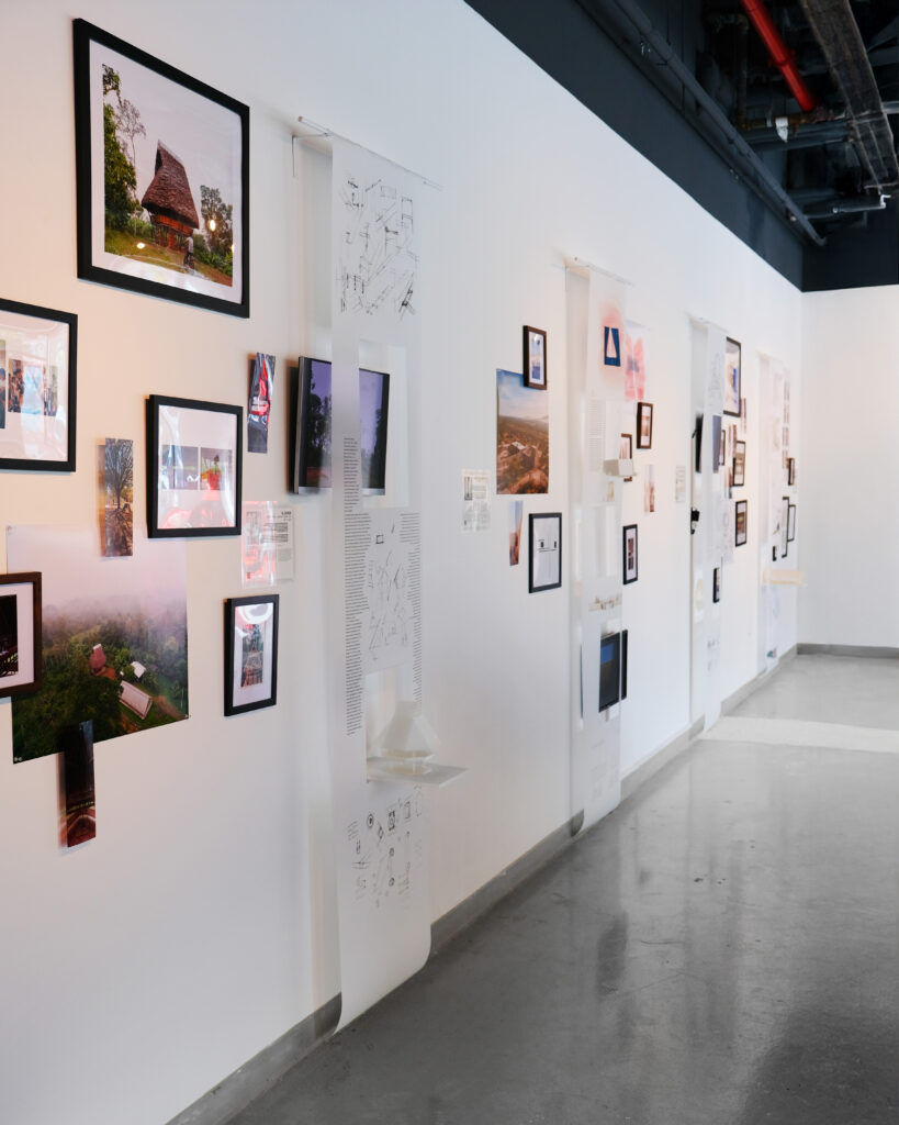 Perspective view down an architectural gallery corridor with polished concrete floors and exposed dark ceiling pipes. The long white exhibition wall features a dynamic multi-media display, including black-framed landscape and architectural photographs, small vertical video monitors, and long white hanging paper banners printed with diagrams, technical drawings, and text. Several wall-mounted mini shelves project outward beneath cutouts in the banners, showcasing small white 3D architectural models.