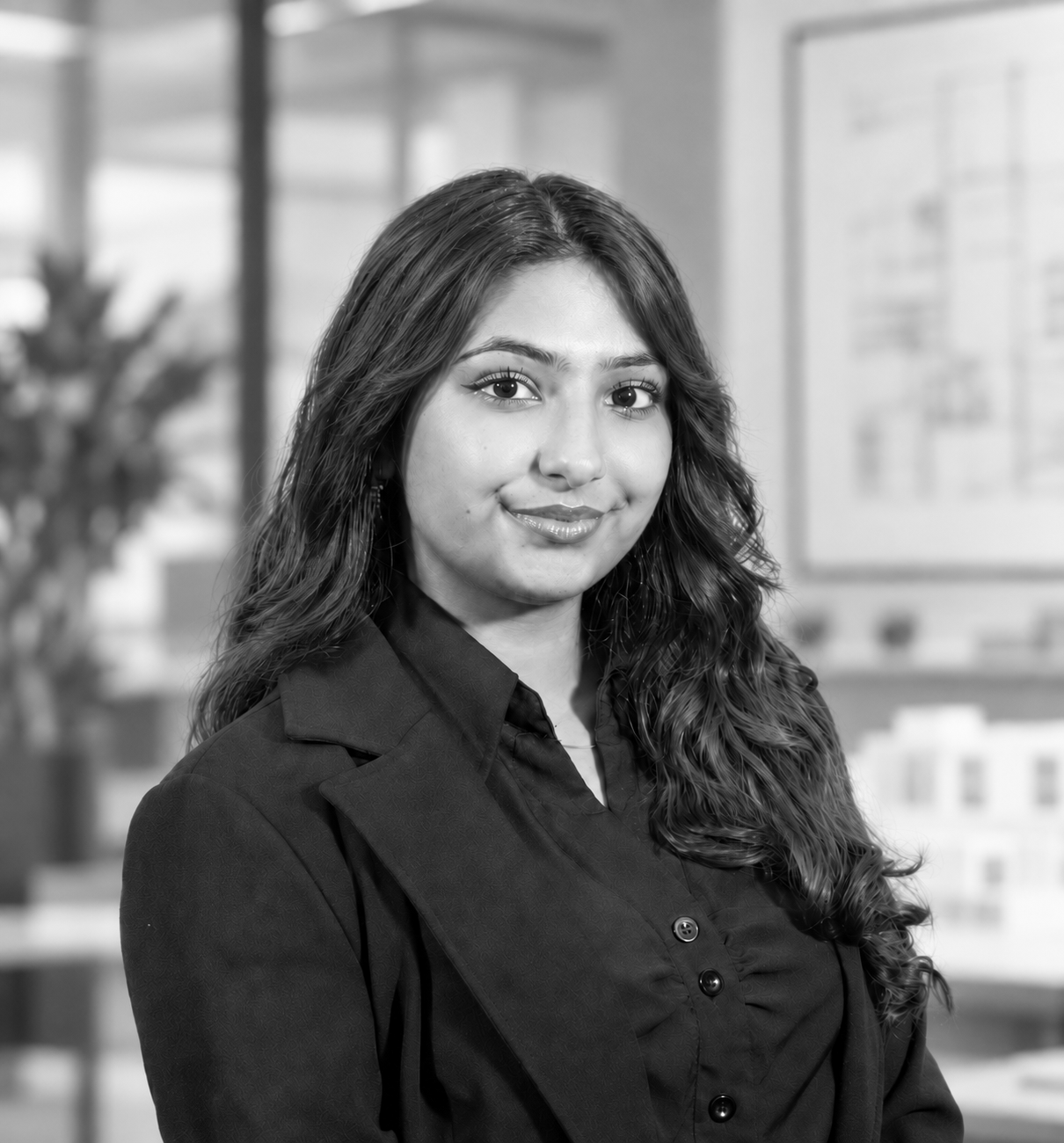 A black and white professional headshot of a young woman with long, dark, wavy hair, wearing a dark button-down shirt and a dark blazer. She is smiling slightly and looking directly at the camera, set against a softly blurred background that suggests an office environment.