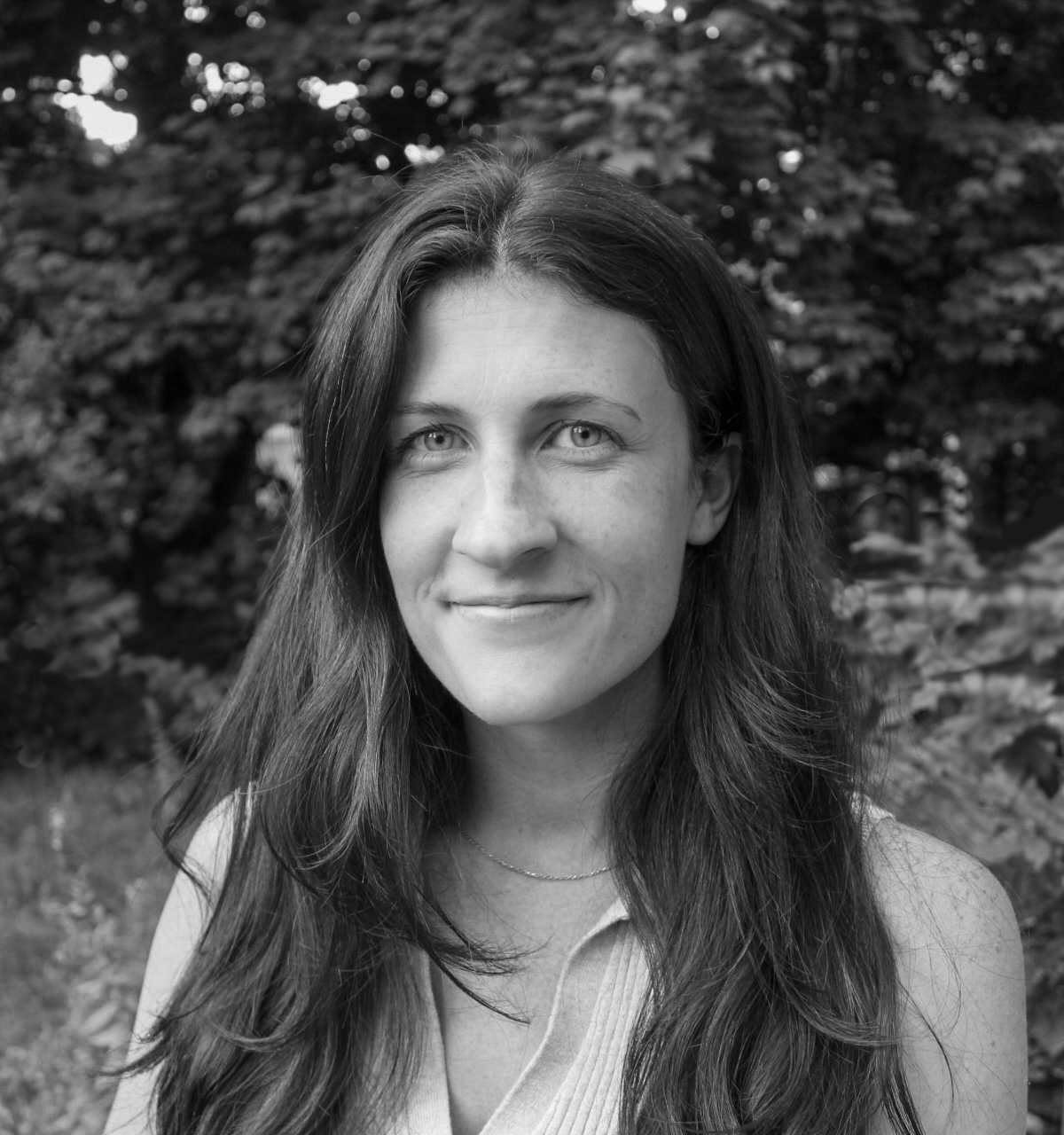Black-and-white headshot of a woman smiling gently at the camera. She has long, dark, center-parted wavy hair resting over her shoulders, wears a light-colored ribbed top and a thin necklace, framed against a softly blurred background of trees and foliage.