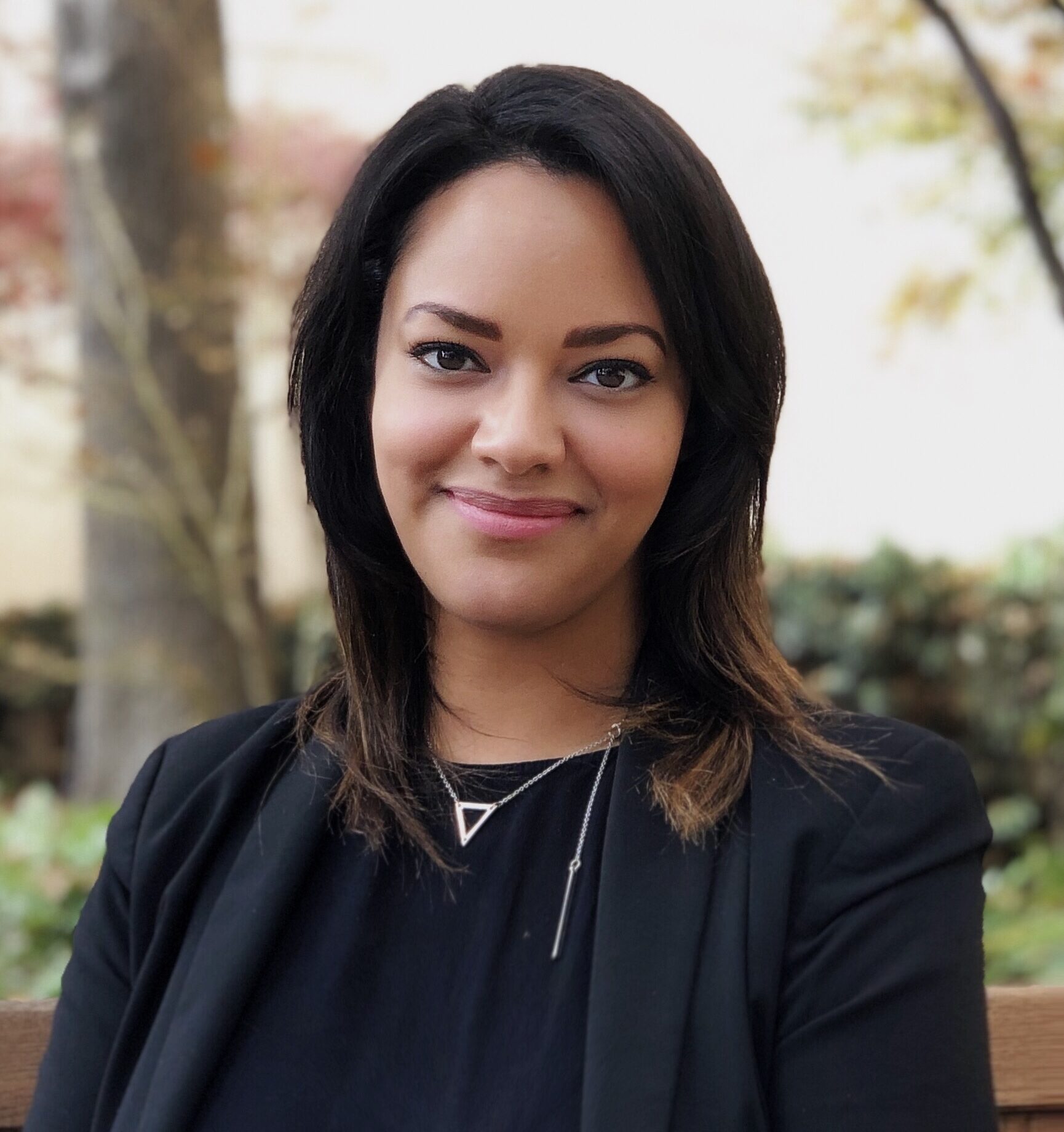 A woman with shoulder-length dark hair and a light smile wearing a black jacket and a silver triangular necklace, posed outdoors against a soft-focus background of trees and foliage.