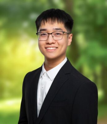 Portrait of a smiling person wearing glasses, a white collared shirt, and a black suit jacket, set against a softly blurred green outdoor background.